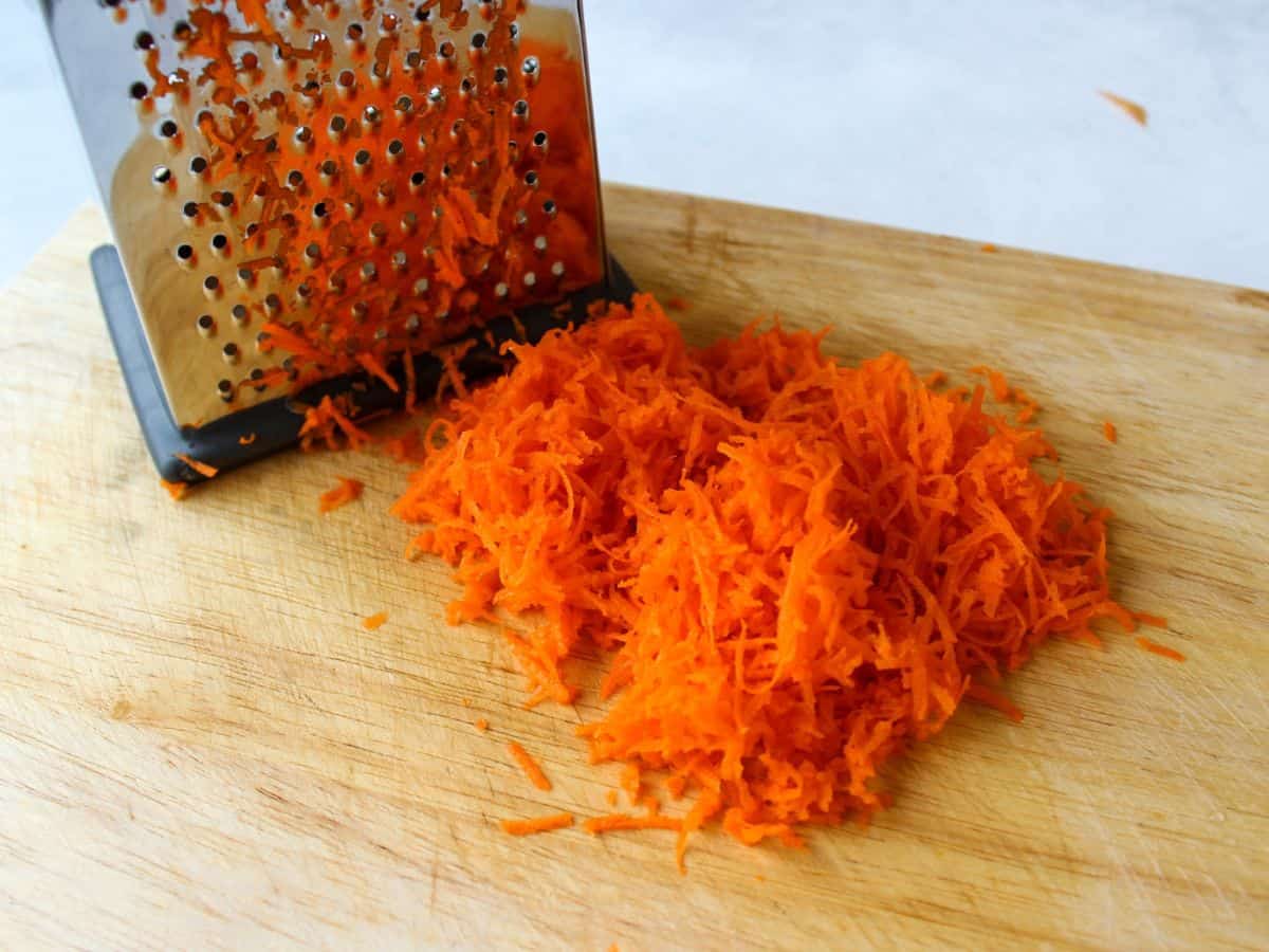 A cutting board with grated carrots and a box grater on the side.