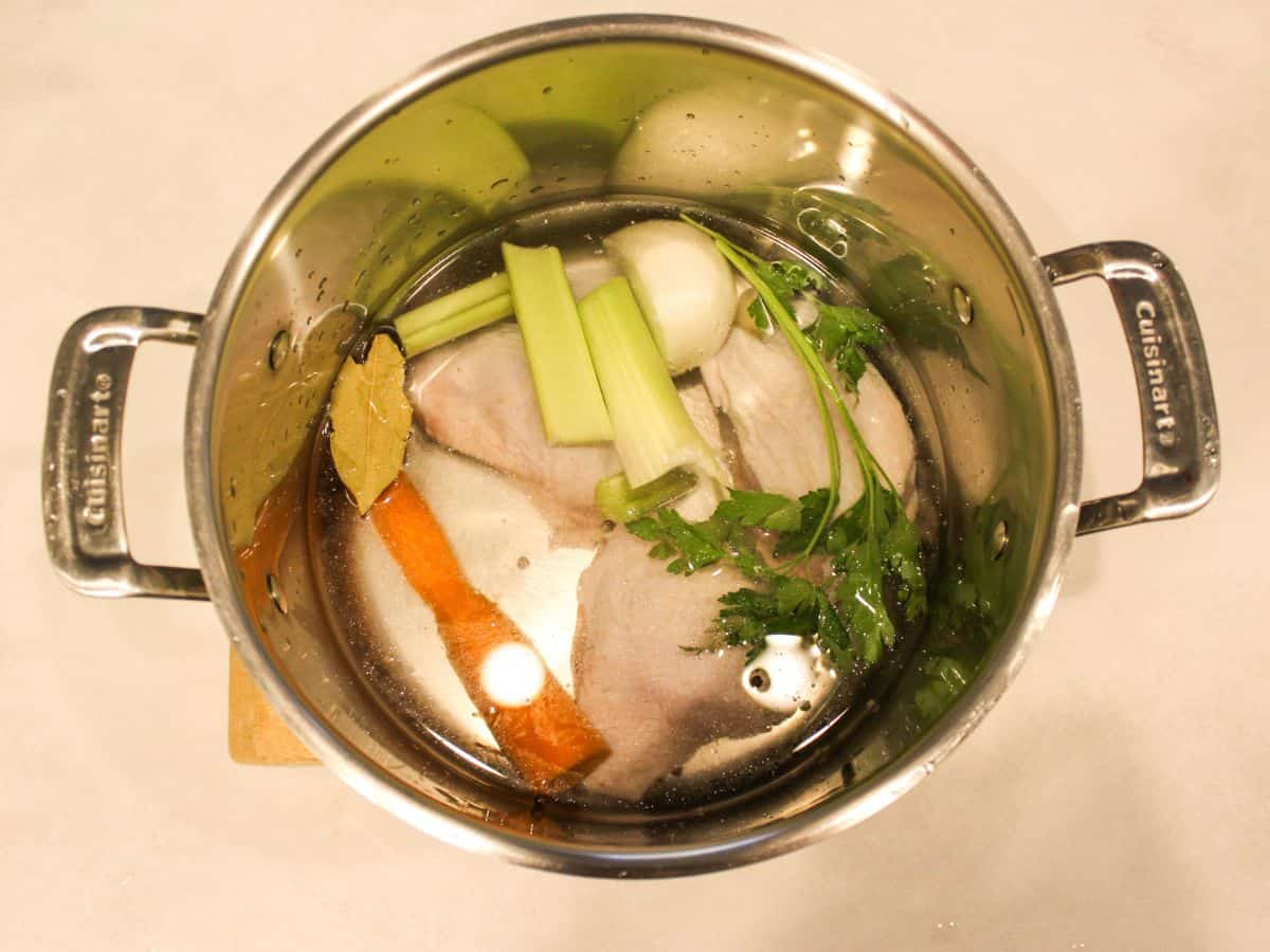 A large stainless steel pot with chicken, celery, carrot, bay leaf, green parsley and water.