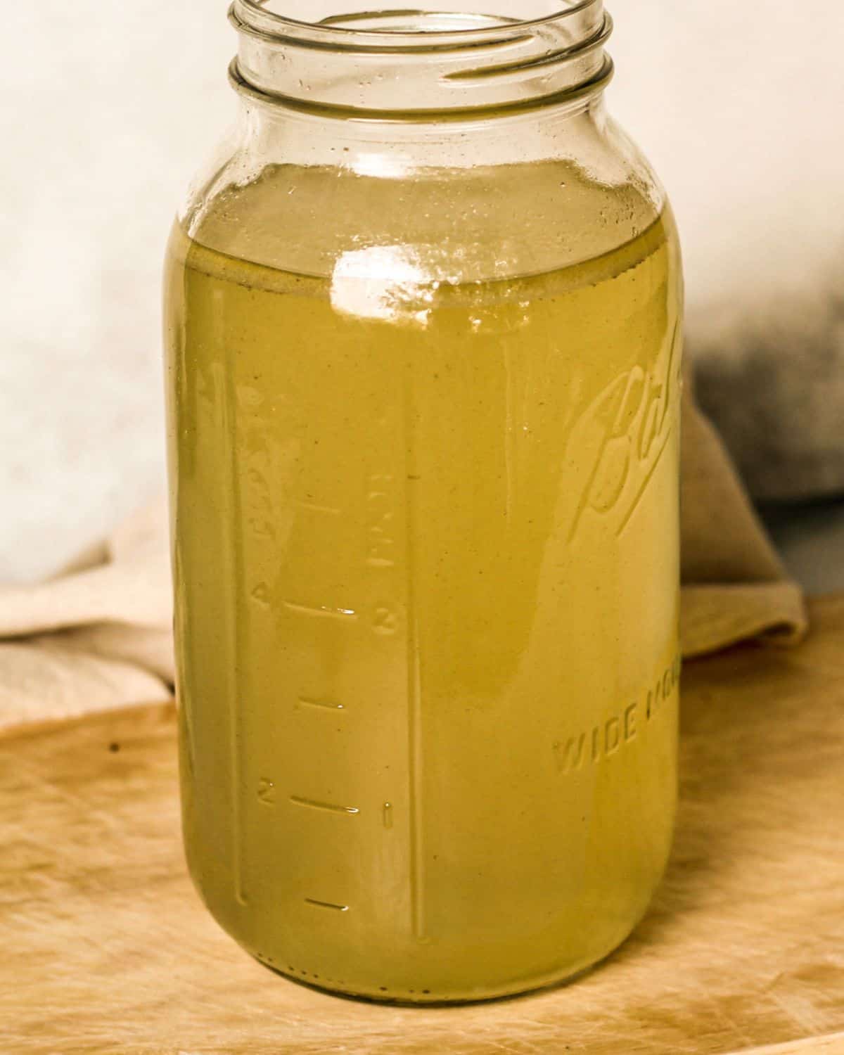 A tall glass jar with yellow broth on a wooden cutting board.