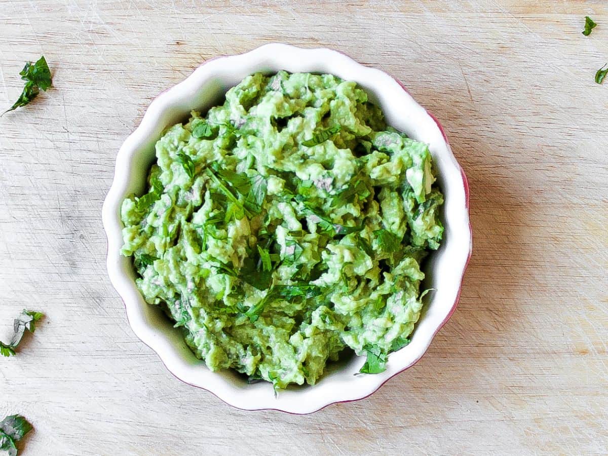 Overhead shot of a bowl with green guacamole. The bowl is on a wooden board. 