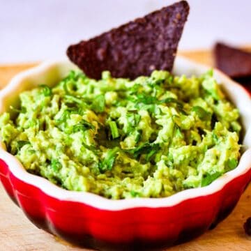 A ceramic red bowl with green chunky guacamole and one black triangle chip dipped into it.