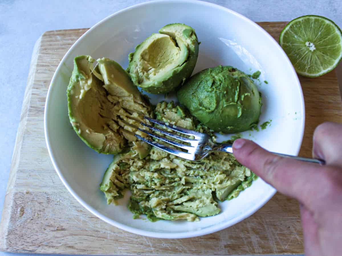 A white large plate on a cutting board with 3 avocado halves and some smashed avocado using a fork.