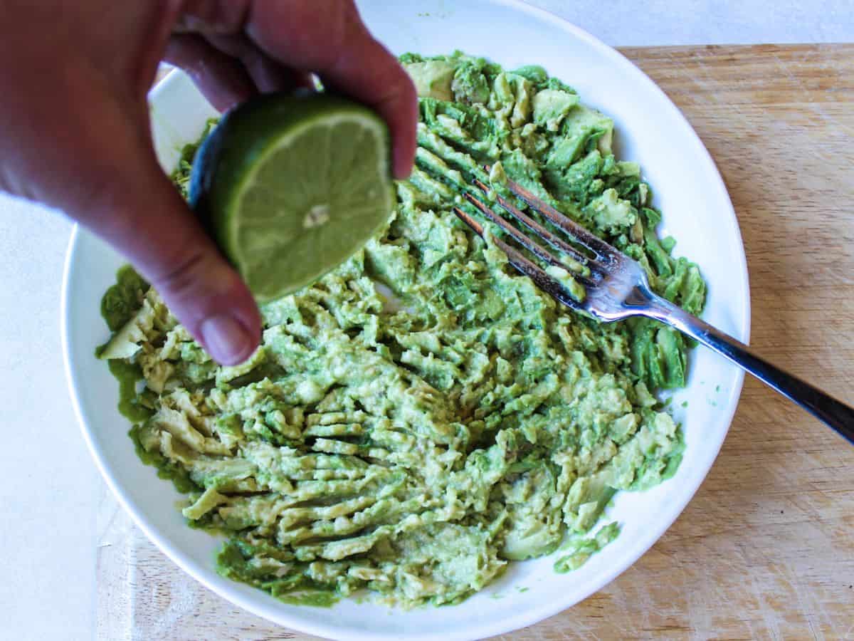 A large white plate with smashed avocado and a hand squeezing the juice out of lime over it.