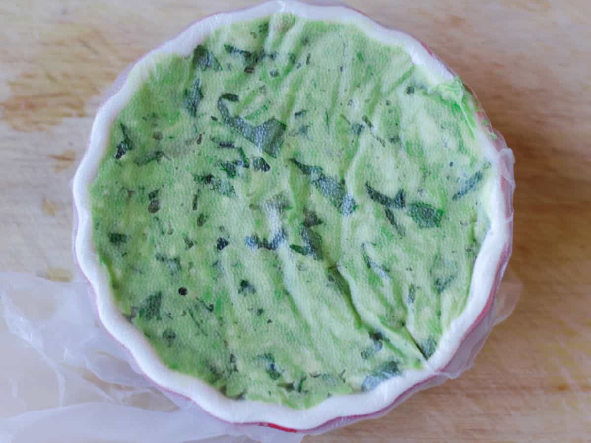 A small round bowl with green dip covered with some plastic wrap on a wooden board.