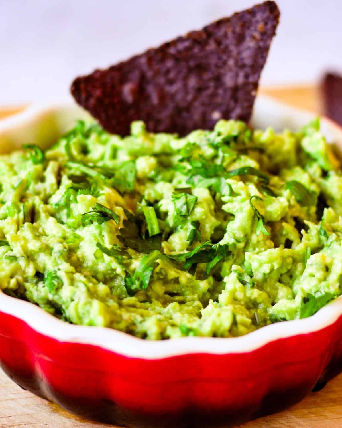 A ceramic red bowl with green chunky guacamole and one black triangle chip dipped into it.