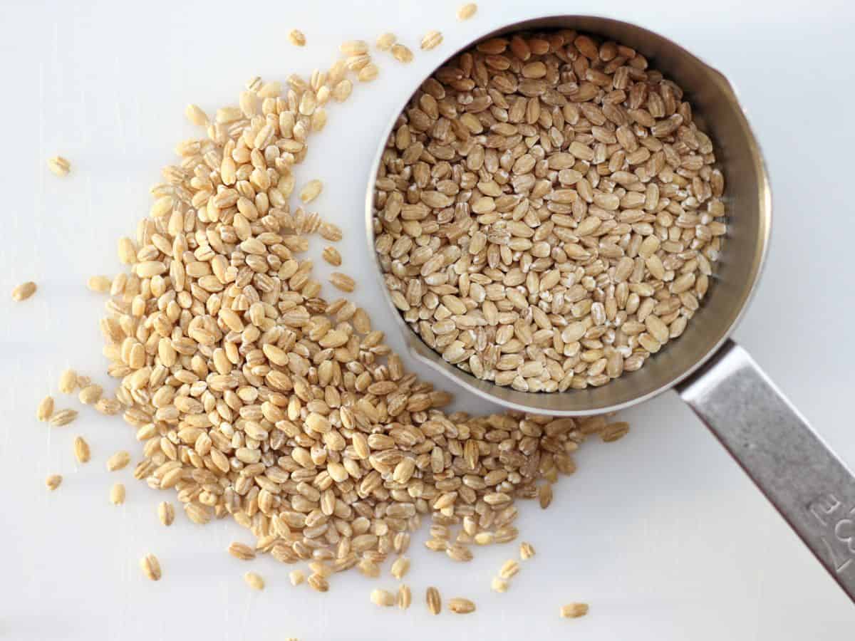 A measuring cup with uncooked barley in it and some grains on the white surface next to the cup.