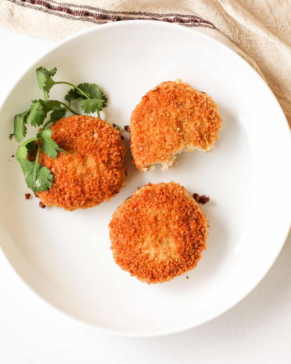 Overhead view with three golden-brown rissoles on a white plate. There is some fresh whole cilantro leaves next on a plate.