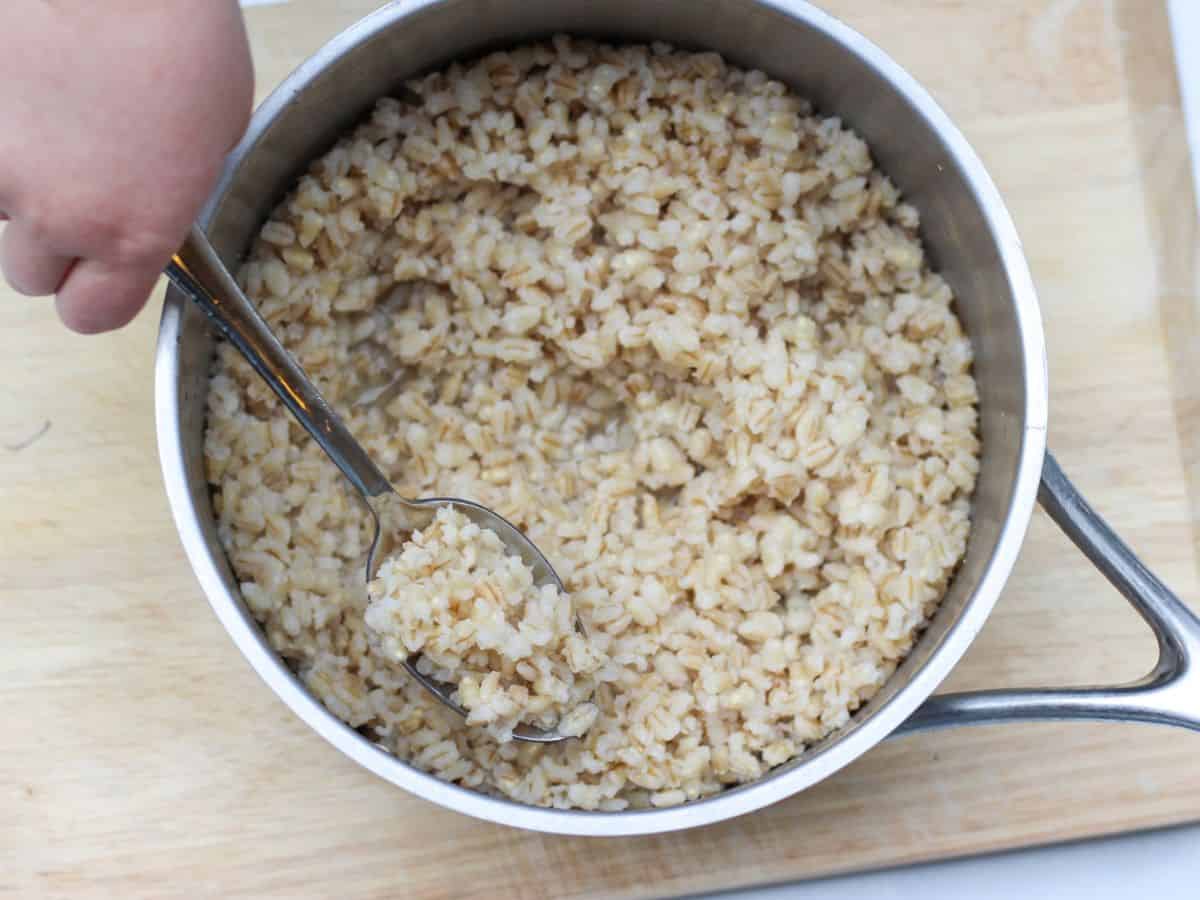 A stainless steel pot on a cutting board. There is cooked barley and a spoon with scooped grain.