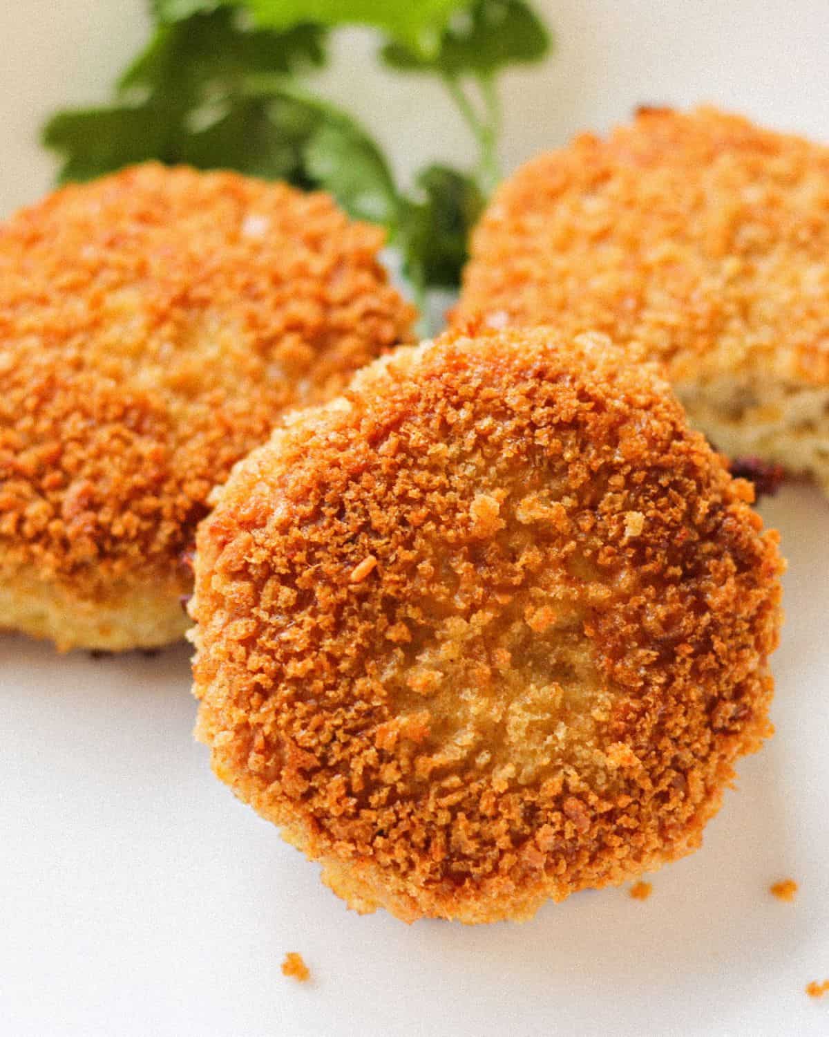 Three breaded cooked patties on a white plate. There is green cilantro in the back.