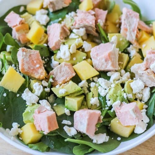 A white bowl with green spinach, cubed avocado, mango, crumbled cheese, and cubes of cooked pink fish.