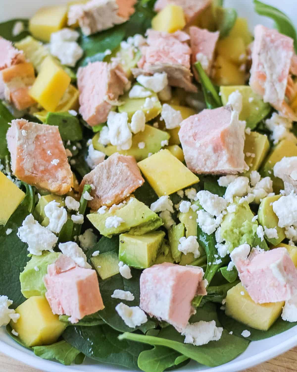 A white bowl with green spinach, cubed avocado, mango, crumbled cheese, and cubes of cooked pink fish.