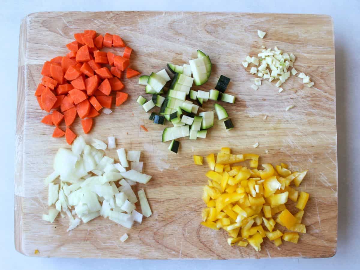 A cutting board with heaps of diced carrots, zucchini, garlic, onion, yellow bell pepper.
