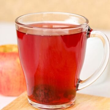 A glass cup filled with red fruit drink. There is an apple in the background.