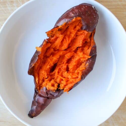 Baked whole sweet potato on a white plate. Some skin on top is removed and the potato flesh is orange and creamy.