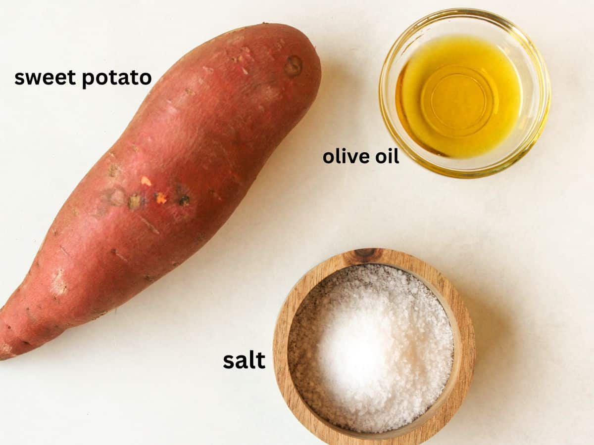 Labeled recipe ingredients on a white surface. One whole sweet potato, small bowl with olive oil and a salt in a round wooden storage box.