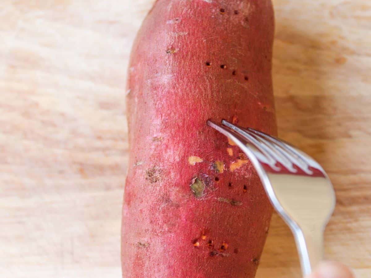 The red skin of a long sweet potato is pricked with a fork.