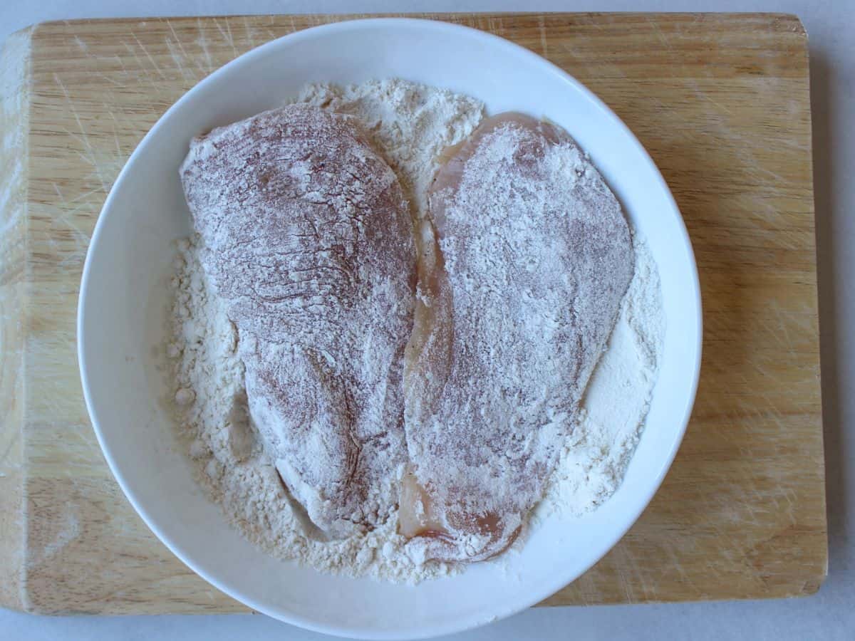 A white shallow dish with flour and 2 coated uncooked chicken breasts.