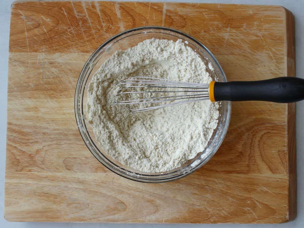 White flour in a glass bowl with a whisk in it on a wooden cutting board.