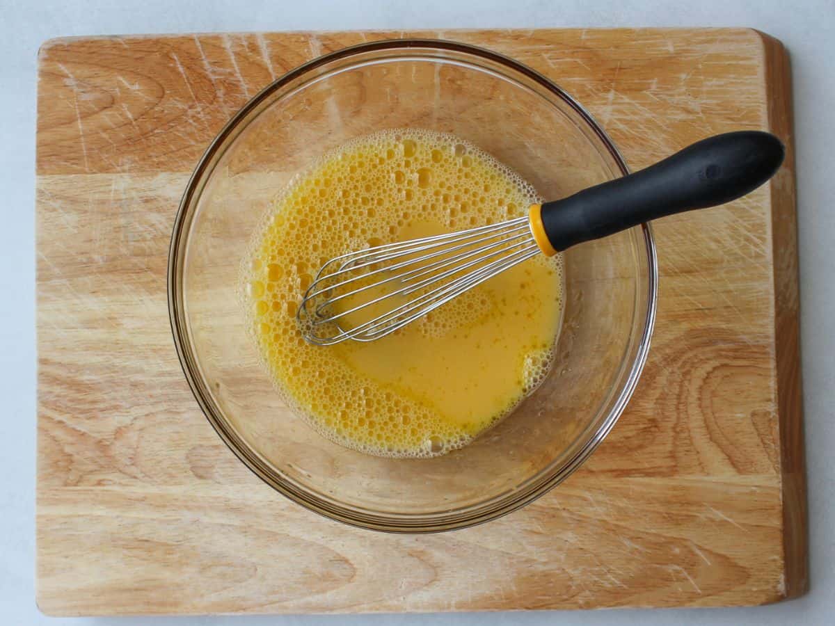 Egg yellow mixture in a glass bowl with a whisk in it on a wooden cutting board.