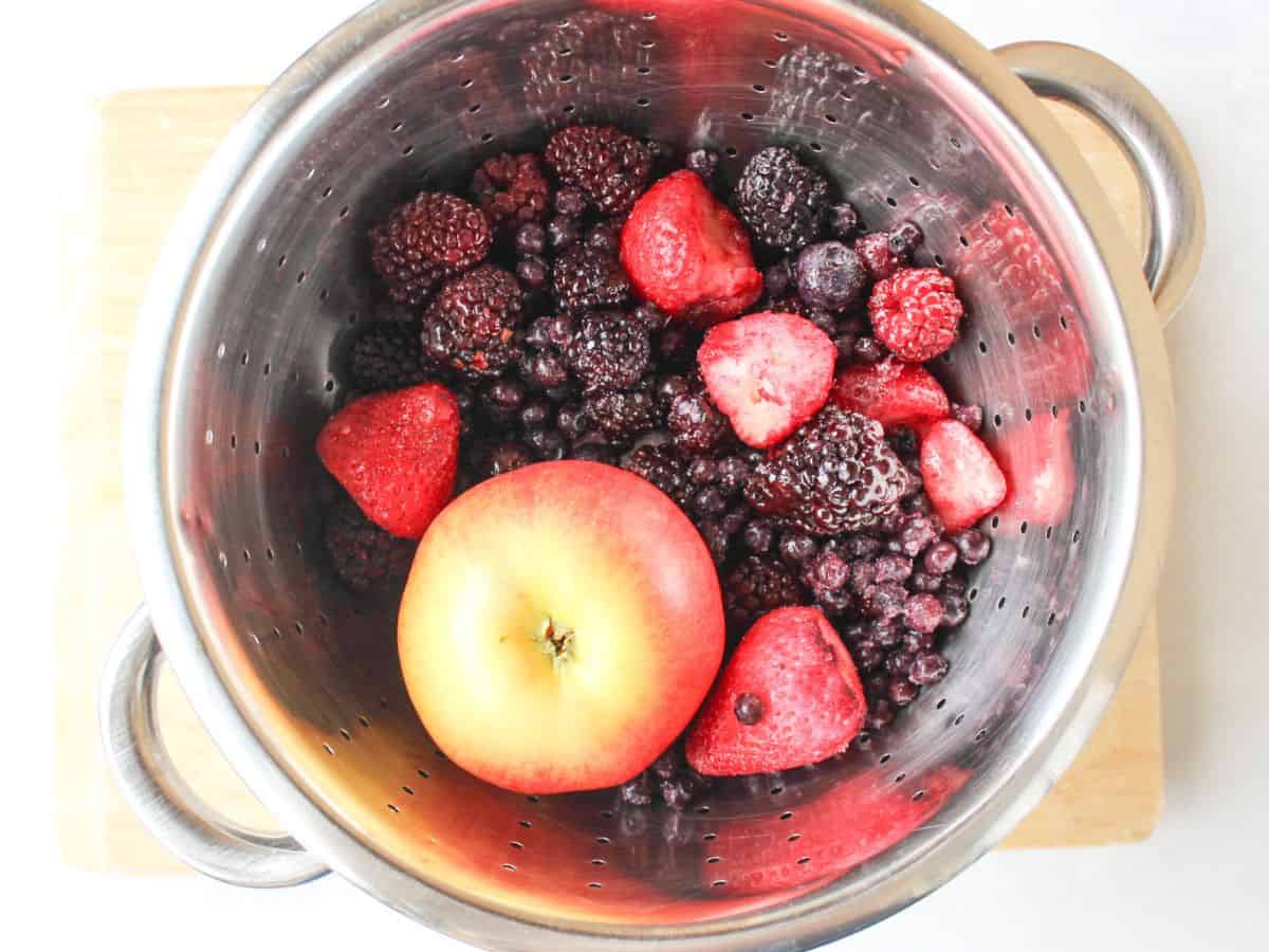 A stainless steel colander with one whole apple, frozen strawberries, frozen blackberries and frozen blueberries.