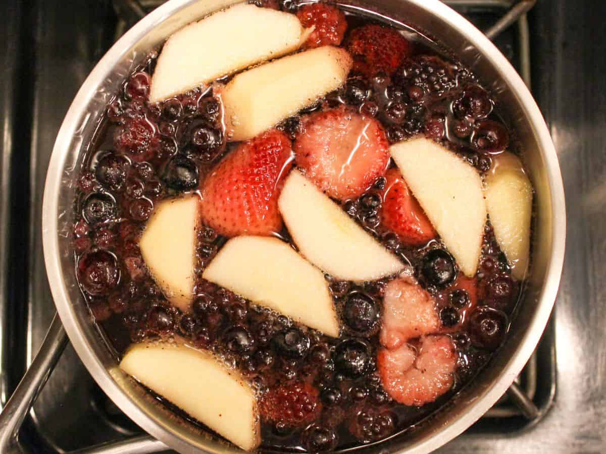 A stainless steel pot with blueberries, strawberries and sliced apples simmering in water.