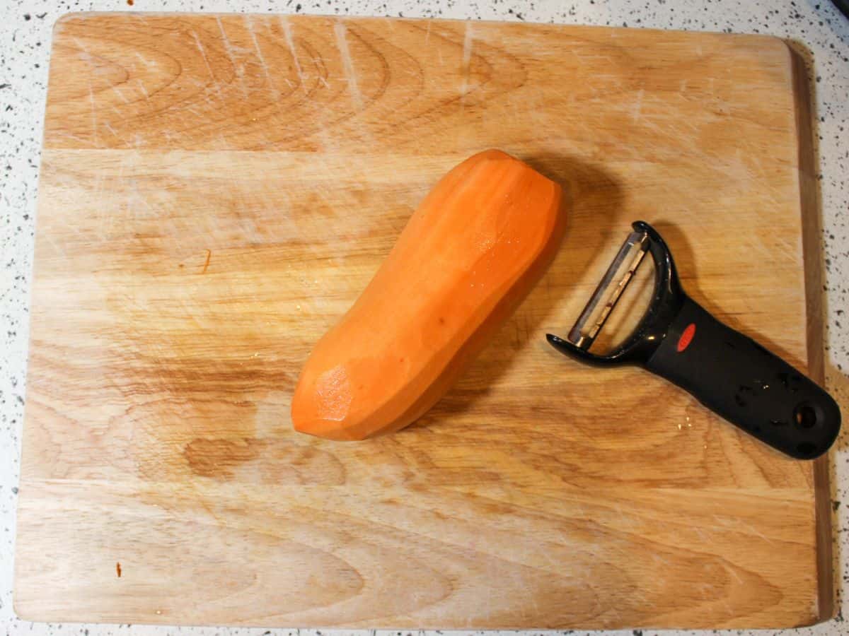 Peeled orange potato on a cutting board with a veggie peeler on the right side. 