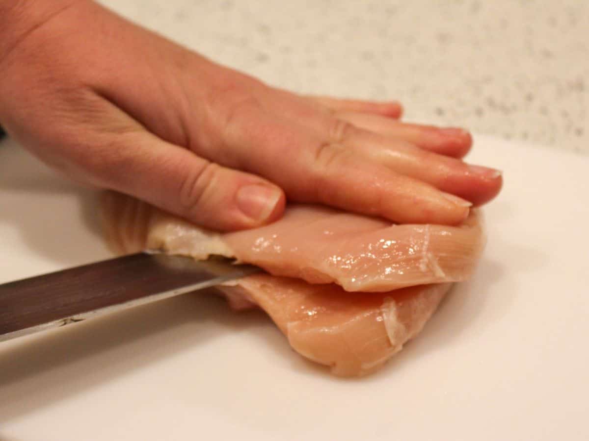 Raw chicken breast being cut into cutlets on a white cutting board with a big knife.