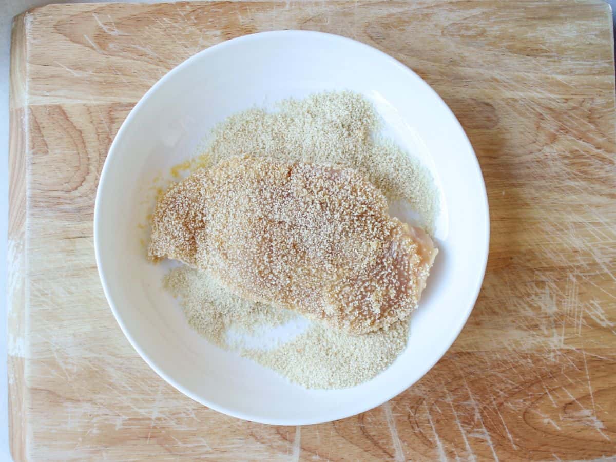 A white dish with bread crumbs and chicken cutlet coated in them.