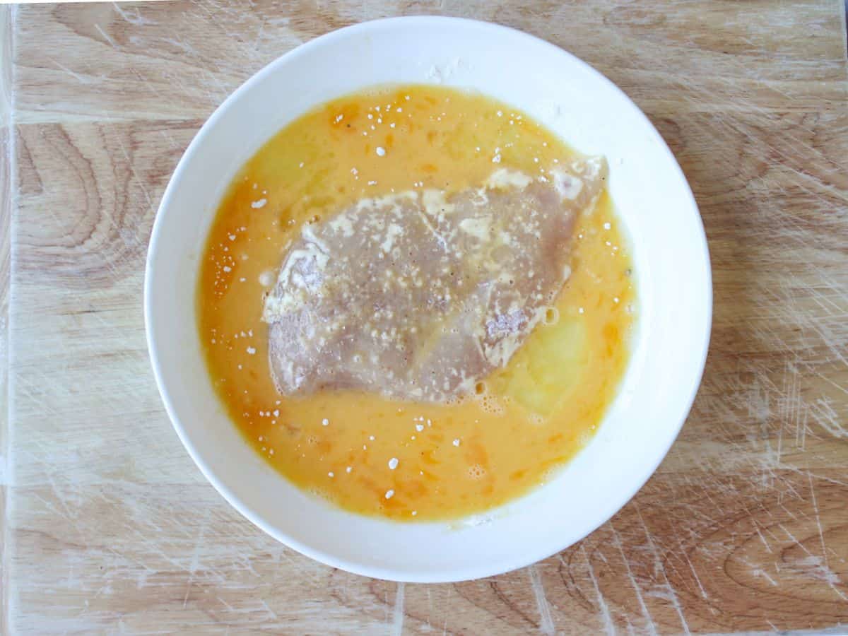 A white dish with yellow egg mixture and chicken cutlet coated in it.