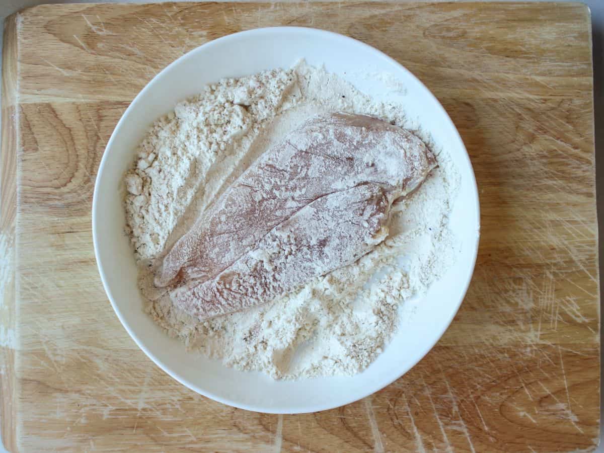 A white dish with flour and chicken cutlet coated in flour.