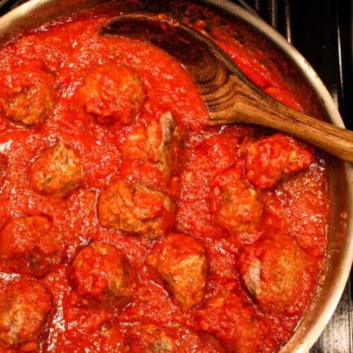 A skillet filled with red tomato sauce and meatballs. There is a wooden serving spoon in the skillet.