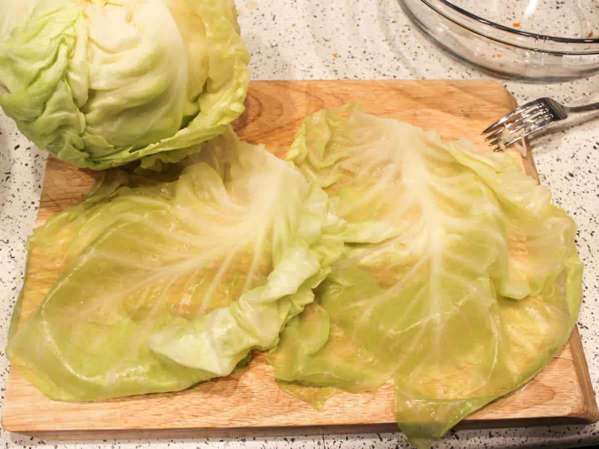 Two green cabbage leaves are laying flat on a cutting board. The head of the cabbage is there on the top left side.