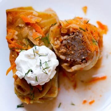 Two cabbage rolls on a white place. One is topped with a dollop of sour cream. The second roll is cut in half and facing the cut side up to show the meat and rice filling. The rolls are sprinkled with fresh chopped green dill on top and there are few shreds of carrot on the plate.