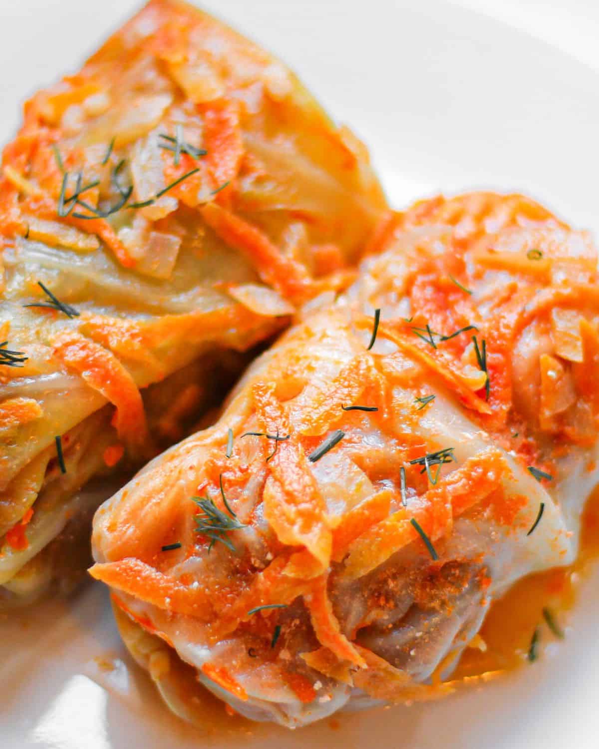 Two cabbage rolls on a white plate are placed next to each other. Green chopped dill is sprinkled on top of the rolls.