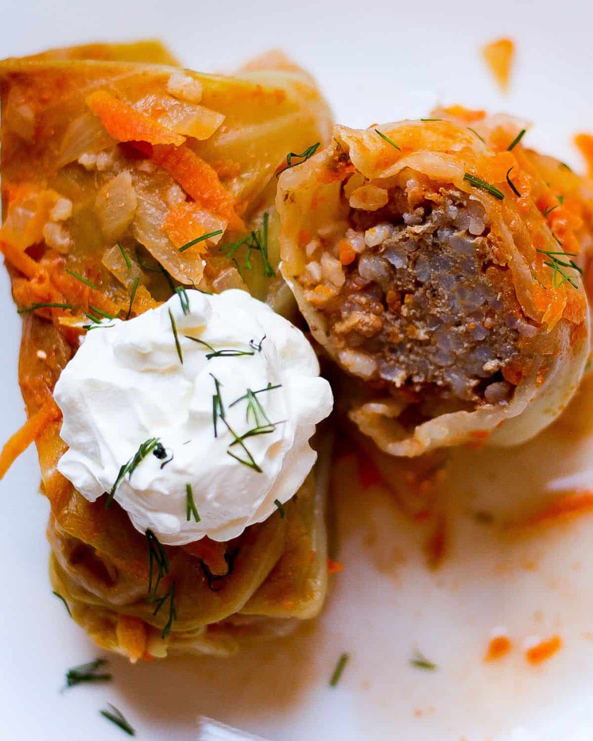 Two cabbage rolls on a white place. One is topped with a dollop of sour cream. The second roll is cut in half and facing the cut side up to show the meat and rice filling. The rolls are sprinkled with fresh chopped green dill on top.