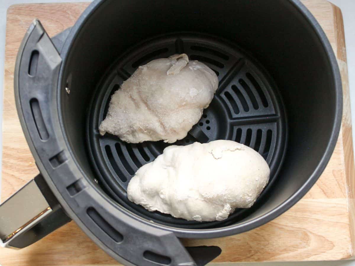 Two unseasoned frozen chicken pieces in a round air fryer basket.