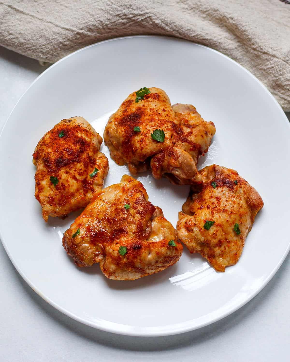 Four cooked boneless skinless chicken thighs in a white plate. The meat is garnished with small pieces of green parsley on top.