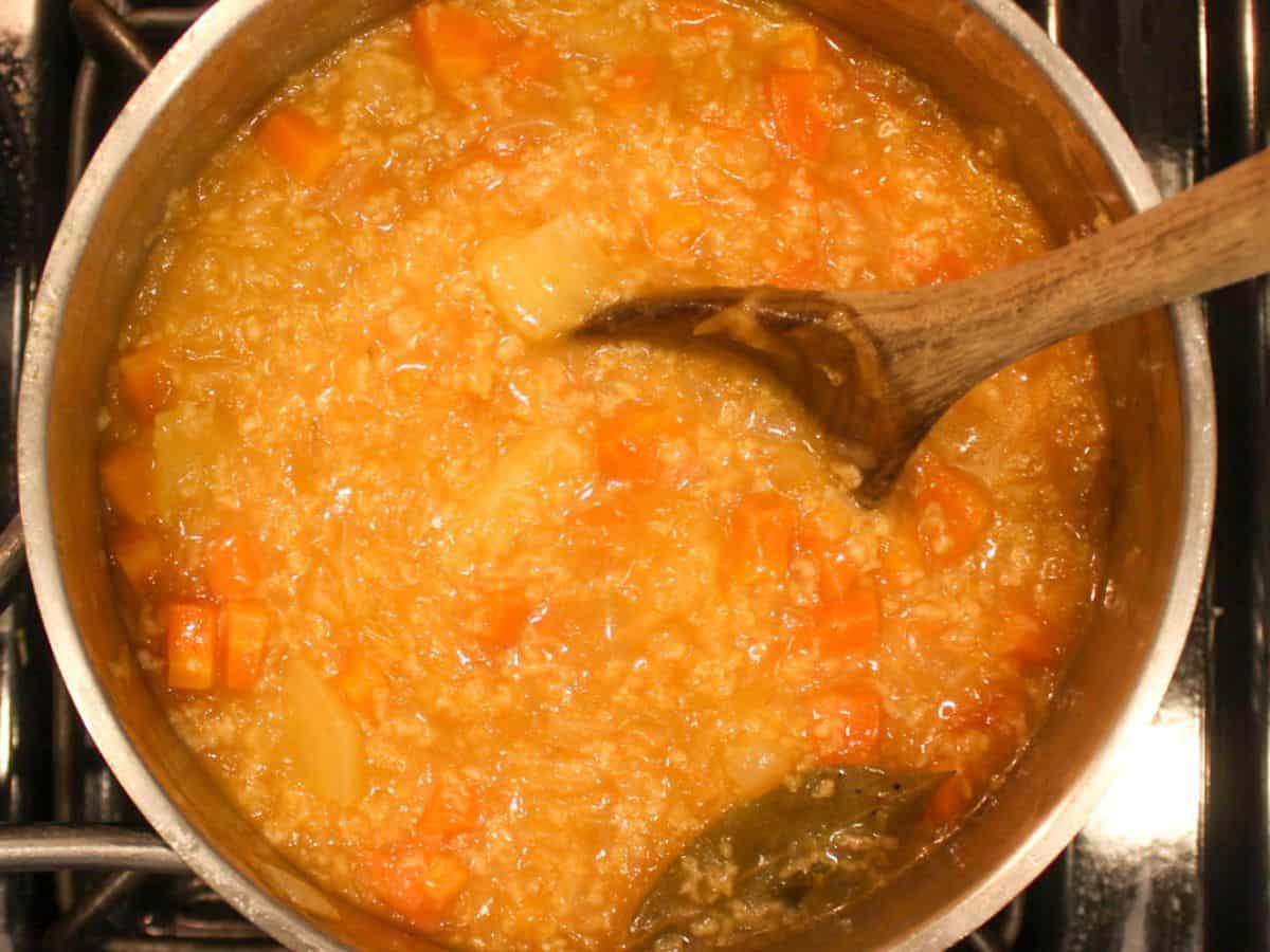 Cabbage soup with diced carrots and one bay leaf in a pot with a wooden spoon in it.