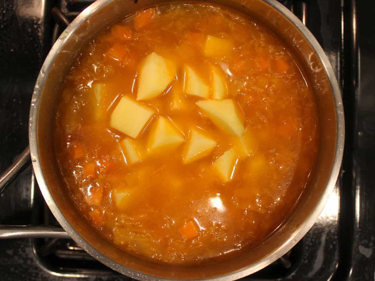 Orange soup with cubed potatoes added in a stainless steel pot.
