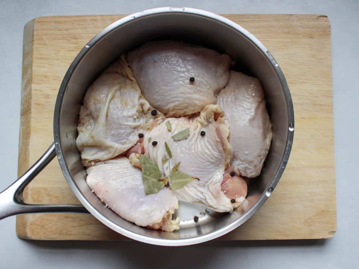 Uncooked chicken thighs in a single layer in a pot with some crushed bay leaves and whole peppercorns.