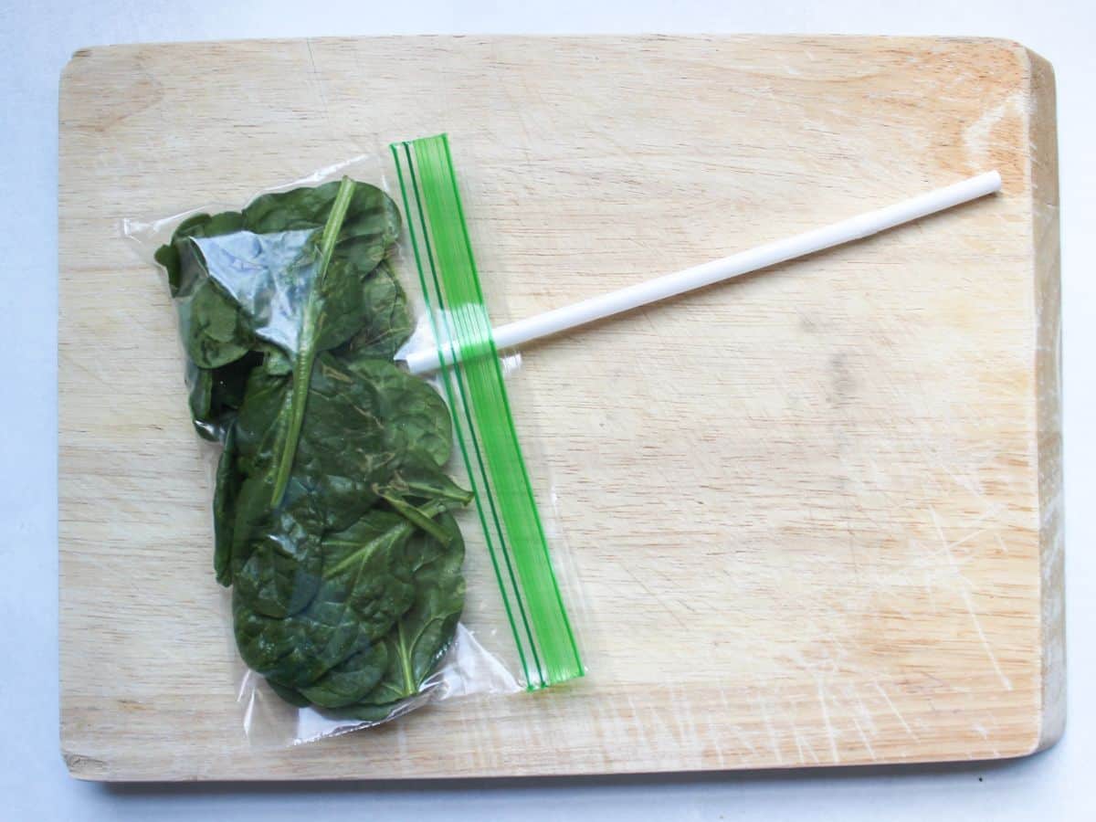A food storage bag with spinach and a drinking straw inserted into the opening to create a vacuum seal.