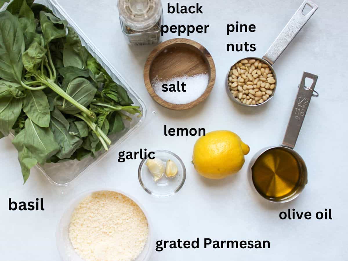 Recipe ingredients on a white surface: fresh basil, black pepper, salt, pine nuts. garlic cloves. whole lemon, olive oil, grated Parmesan cheese.