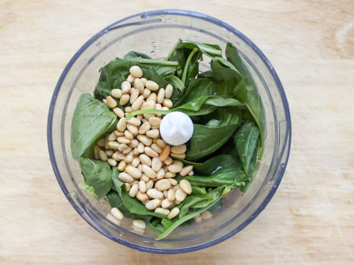 Green basil leaves and pine nuts in a clear food processor container.