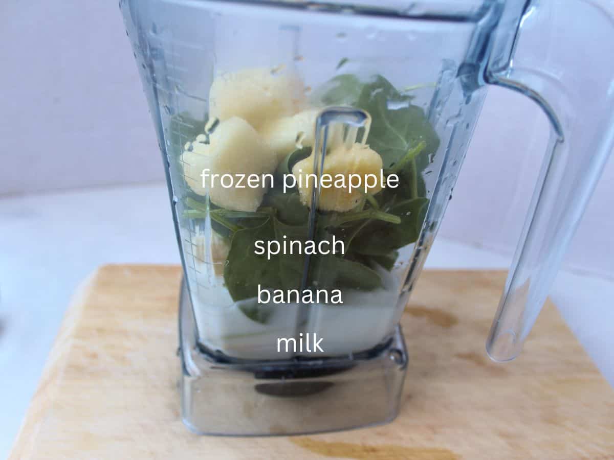 A blender container with milk, spinach, and chunks of frozen pineapple.