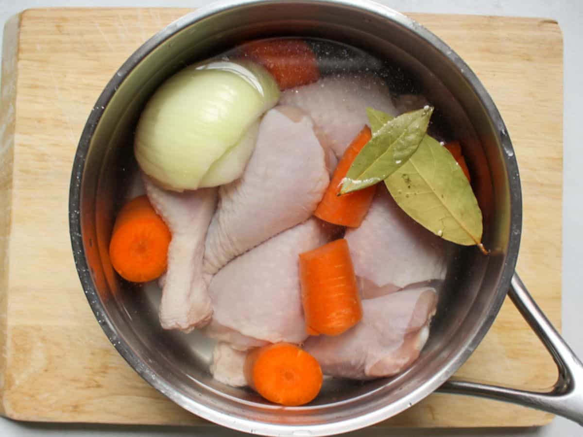 Raw chicken legs, carrots, onion and bay leaf in a pot filled with water.