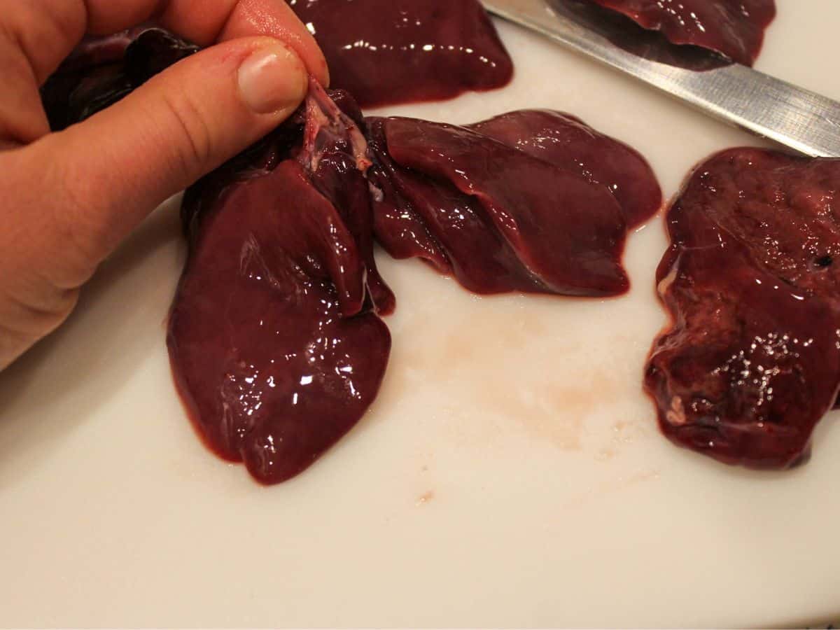 Raw chicken liver on a cutting board being held by the connecting tissue that should be trimmed.