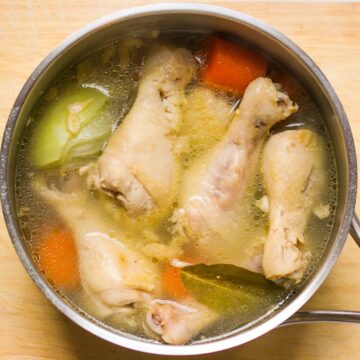 Boiled chicken legs in a broth with carrots, onions and bay leaf in a stainless steel pot.