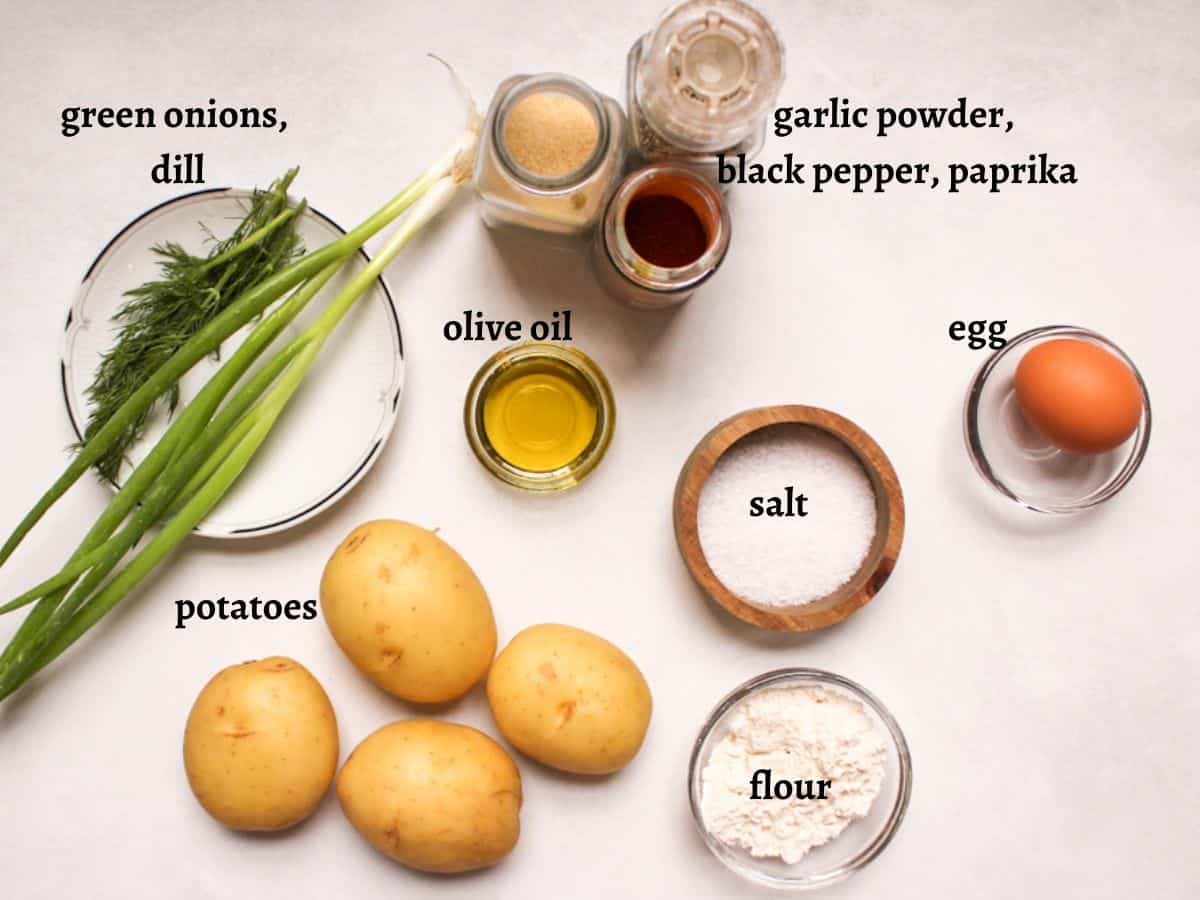 Recipe ingredients on a white surface: green onions, fresh dill, 4 potatoes, olive oil, garlic powder, paprika, black pepper, salt, egg, flour.