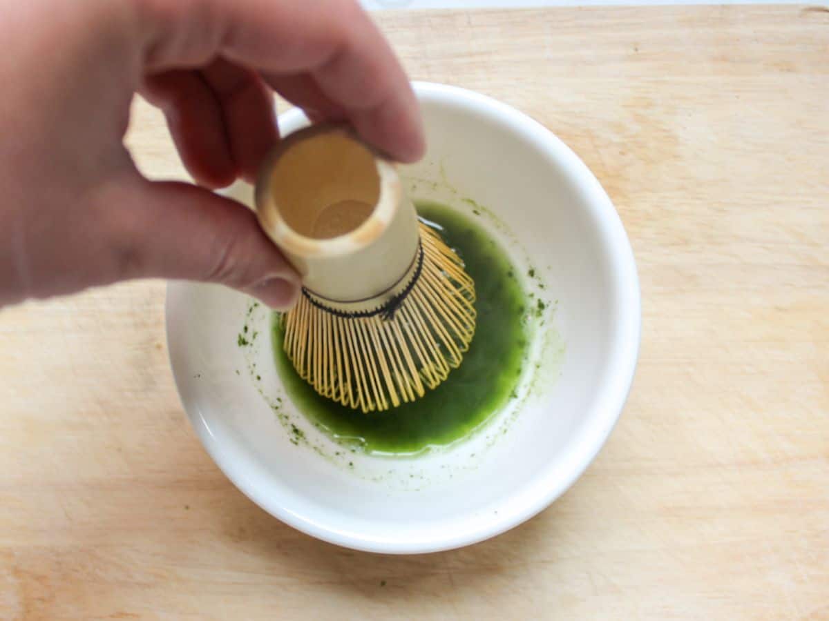 Matcha powder and hot water mixed with a bamboo whisk in a white bowl.