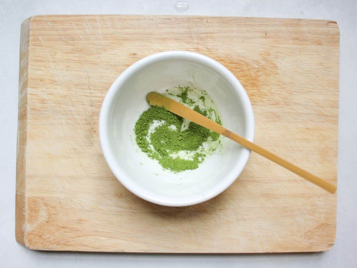Green matcha powder in a white bowl with a bamboo measuring stick in it.
